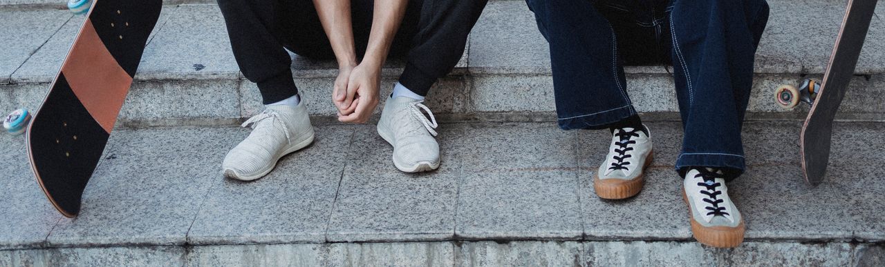 Crop faceless friends in trendy sneakers resting on stone stairs in city with skateboards